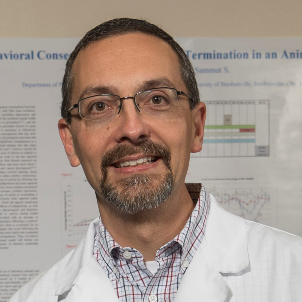 Close-up of a middle-aged man with glasses and a goatee wearing a white lab coat, standing in front of science posters.