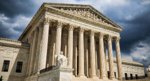 Front view of a neoclassical courthouse with tall marble columns, statues, and a dramatic cloudy sky behind it.