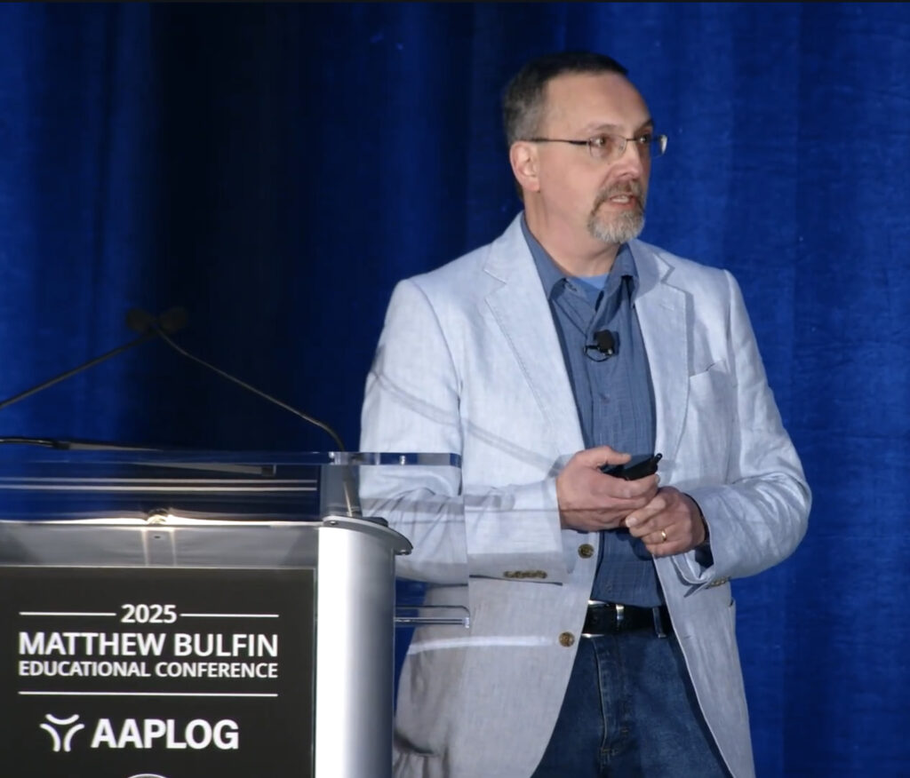 Man in a light blazer and glasses speaking at a podium on a blue-curtained stage; signage shows '2025 Matthew Bulfin Educational Conference, AAPLOG'.