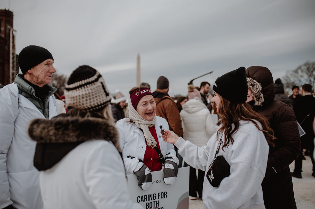 Older woman in a white coat being interviewed at a winter outdoor event; reporter holds a microphone while the crowd gathers, with the Washington Monument in the distance.