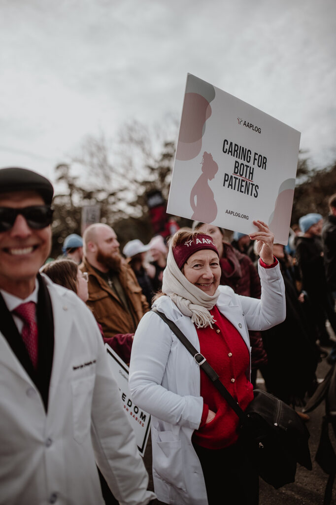 Woman in a white coat and red sweater holding a sign reading 'Caring for Both Patients' at a protest; crowd in the background.