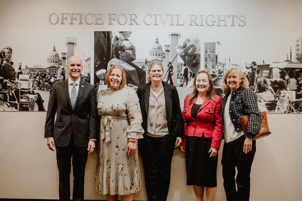 Five professionally dressed individuals smile for a group photo in front of a mural reading 'Office for Civil Rights'.