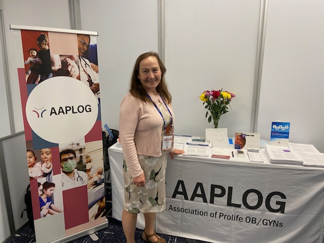 Woman standing beside an AAPLOG exhibition booth with a banner, table of pamphlets, and a vase of flowers at a conference.