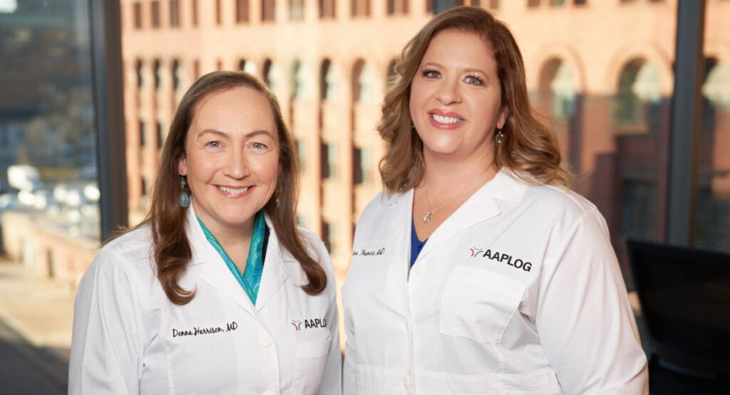 Two female doctors in white coats stand side by side in a bright office, smiling at the camera; coats show the AAPLOG logo.