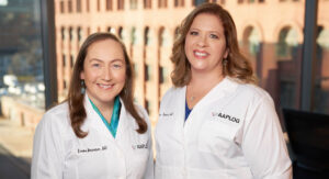 Two female doctors in white coats stand side by side in a bright office, smiling at the camera; coats show the AAPLOG logo.
