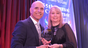 Two adults on stage smiling as they hold a glass award together, with a blue banner in the background.
