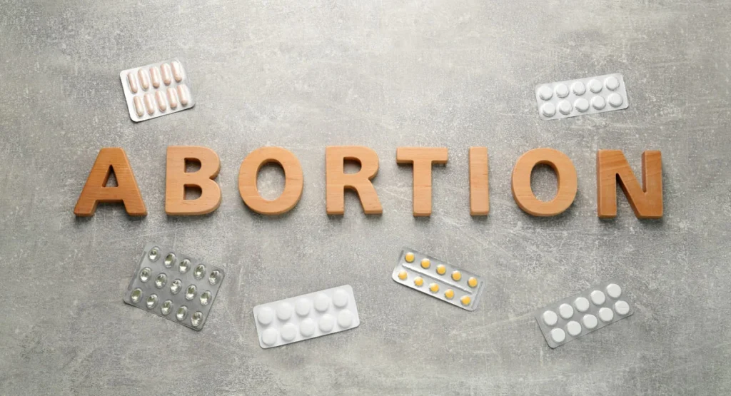 Wooden letters spell ABORTION on a gray surface, surrounded by blister packs of pills.