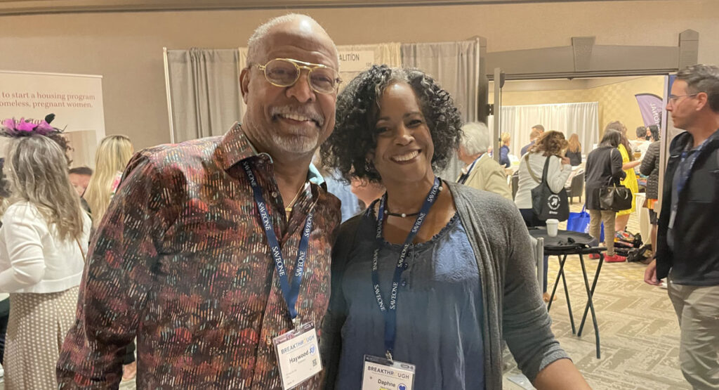 Two smiling attendees wearing conference badges pose for a photo at a busy expo with booths in the background.